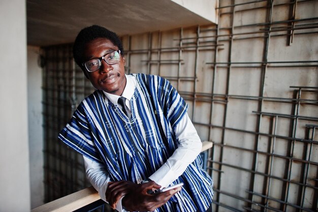 Hombre de negocios africano con ropa y gafas tradicionales con teléfono móvil a mano posado en un edificio moderno interior