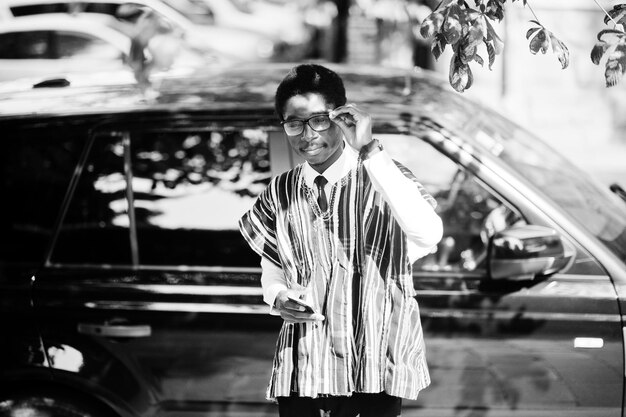 Hombre de negocios africano con ropa tradicional y anteojos con teléfono móvil contra suv de coche negro Gente africana rica