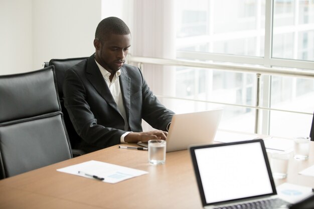 Hombre de negocios africano serio que trabaja en el ordenador portátil que se sienta en la mesa de reuniones