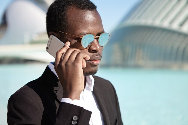 Hombre de negocios afroamericano confiado con traje formal negro y lentes de espejo redondos revisando el correo de voz en su camino de regreso a la oficina después del almuerzo