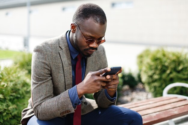 Hombre de negocios afroamericano trabaja en su teléfono inteligente sentado en el banco fuera