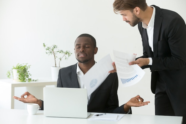 Hombre de negocios afroamericano tranquilo practicando yoga en el trabajo ignorando jefe enojado