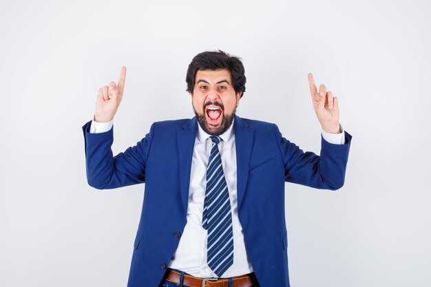 Hombre de negocios apuntando hacia arriba con los dedos índices en traje formal y mirando feliz, vista frontal.