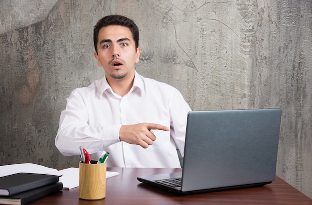 Hombre de negocios apuntando a la computadora portátil y sentado en el escritorio. Foto de alta calidad