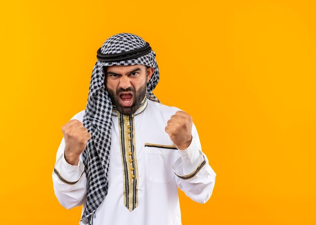 Hombre de negocios árabe en ropa tradicional con cara enojada apretando los puños de pie sobre la pared naranja