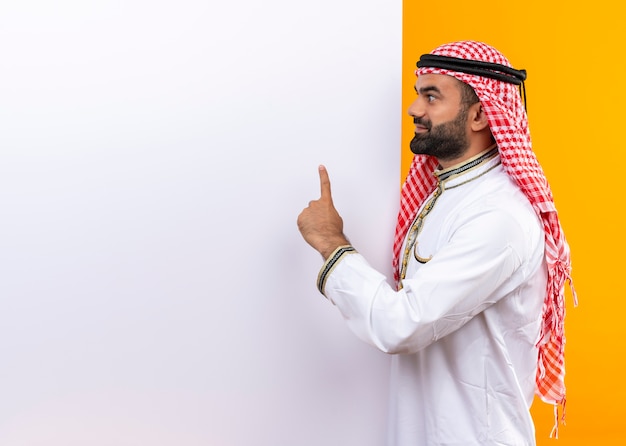 Hombre de negocios árabe en ropa tradicional con cartelera en blanco blanco apuntando con el dedo hacia arriba sonriendo de pie sobre la pared naranja