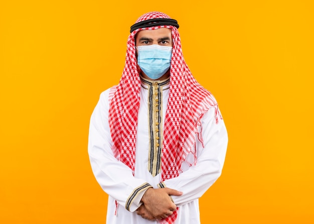 Foto gratuita hombre de negocios árabe en ropa tradicional y máscara protectora facial con expresión de confianza de pie sobre la pared naranja