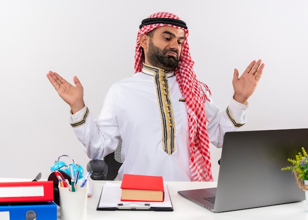 Hombre de negocios árabe en ropa tradicional sentado en la mesa con computadora portátil mirando confundido y desorientado levantando los brazos sin respuesta trabajando en la oficina