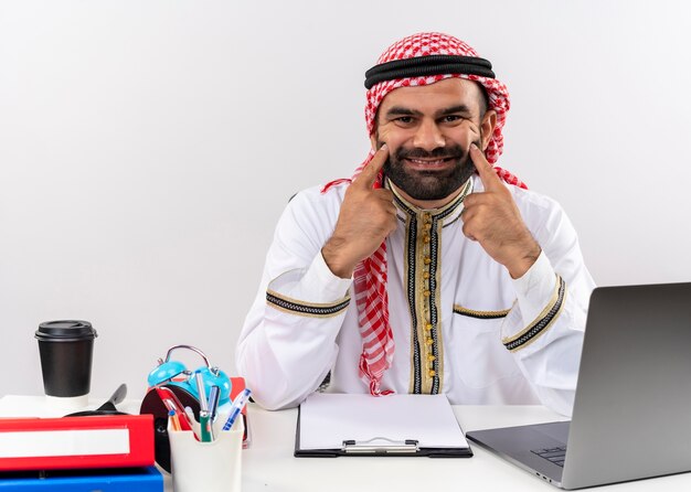 Hombre de negocios árabe en ropa tradicional sentado en la mesa con ordenador portátil apuntando con los dedos a su sonrisa sonrisa falsa trabajando en la oficina