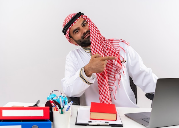 Hombre de negocios árabe en ropa tradicional sentado en la mesa con ordenador portátil sonriendo confiado apuntando con el dedo hacia el lado trabajando en la oficina