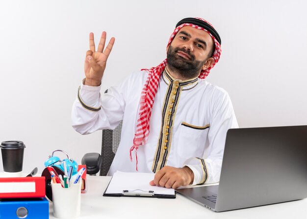 Hombre de negocios árabe en ropa tradicional sentado en la mesa con ordenador portátil sonriendo mostrando y apuntando hacia arriba con los dedos número tres trabajando en la oficina