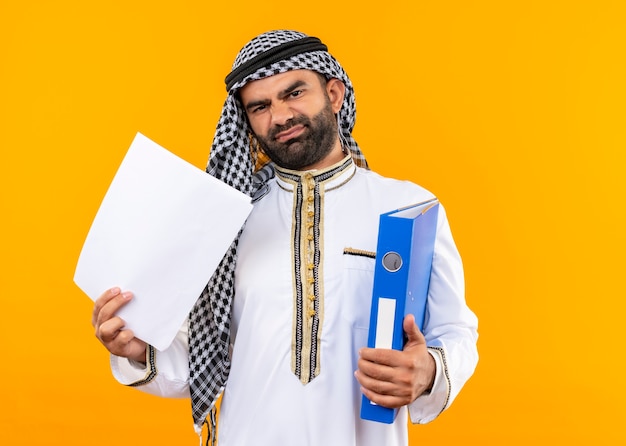 Hombre de negocios árabe en ropa tradicional sosteniendo una carpeta y páginas en blanco disgustado de pie sobre una pared naranja