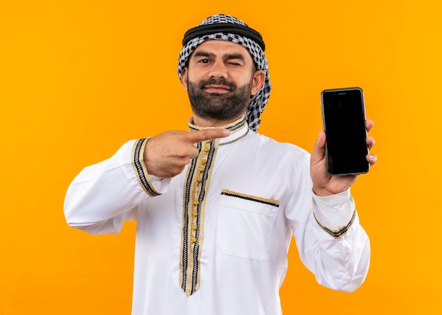 Foto gratuita hombre de negocios árabe en ropa tradicional sosteniendo el teléfono inteligente apuntando con el dedo hacia él mirando confiado guiño de pie sobre la pared naranja