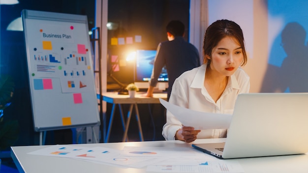 Hombre de negocios asiático apaga la computadora y se despide de su colega que todavía trabaja cuando sale del trabajo después de terminar de trabajar horas extras en la noche de la pequeña oficina en casa moderna. Concepto de asociación de compañeros de trabajo.