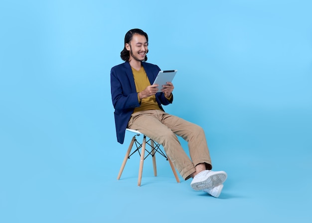 Hombre de negocios asiático feliz sonriendo usando una tableta digital mientras está sentado en una silla en azul brillante.
