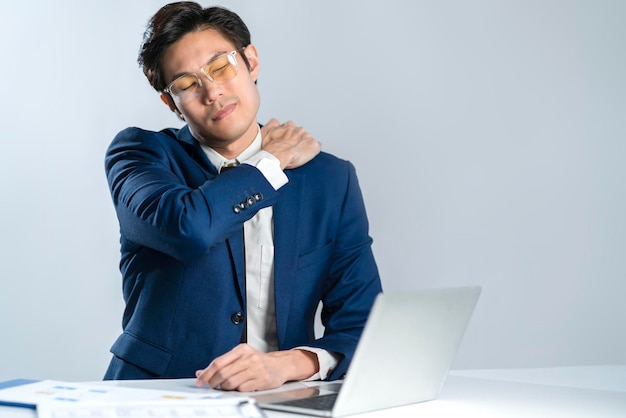 Un hombre de negocios asiático usa un traje formal sentado en la oficina sintiendo dolor corporal por el síndrome de la oficina, varios síntomas, incluida la inflamación del cuello, el hombro y los músculos de la espalda, tiro del estudio, fondo blanco.