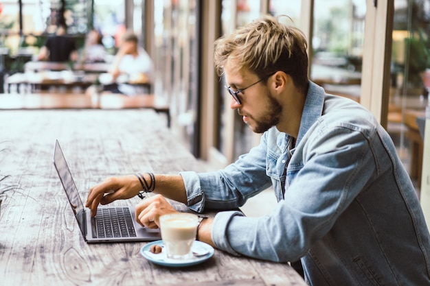 El hombre de negocios atractivo joven en un café trabaja para una computadora portátil, bebe café.