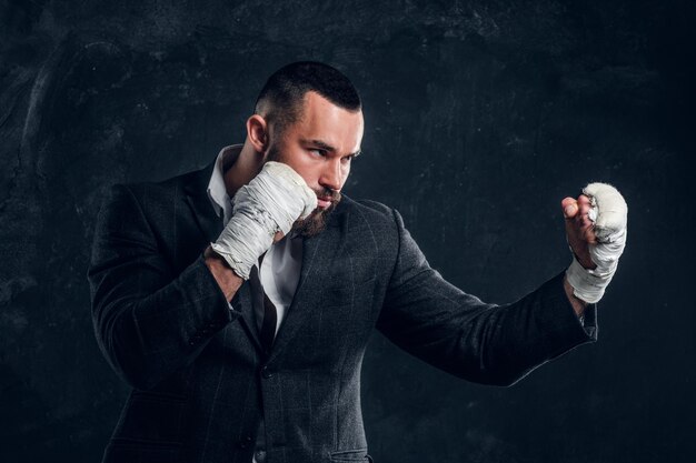 Un hombre de negocios barbudo enojado con traje y guantes protectores de kickboxing está posando para un fotógrafo en un estudio fotográfico oscuro.