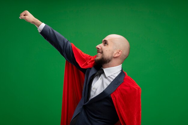 Hombre de negocios barbudo de superhéroe en capa roja haciendo gesto ganador con la mano sonriendo seguro listo para ayudar a pararse sobre la pared verde