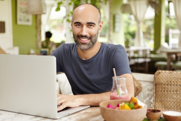Hombre de negocios barbudo vestido casualmente revisando el correo electrónico en su computadora portátil durante el desayuno, sentado en un café agradable, bebiendo batido con expresión de cara feliz y segura, disfrutando de las vacaciones