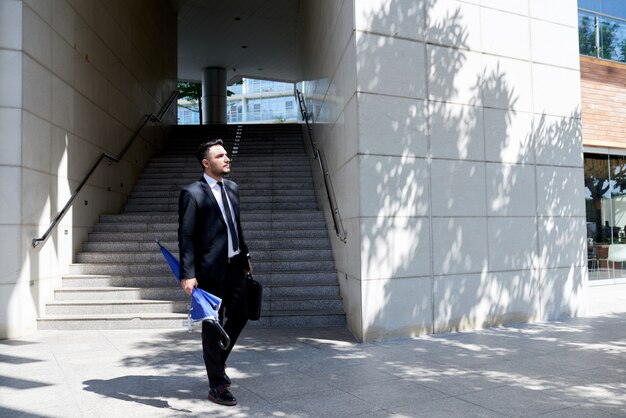 Hombre de negocios caucásico en traje con paraguas caminando delante de la oficina y la escalera