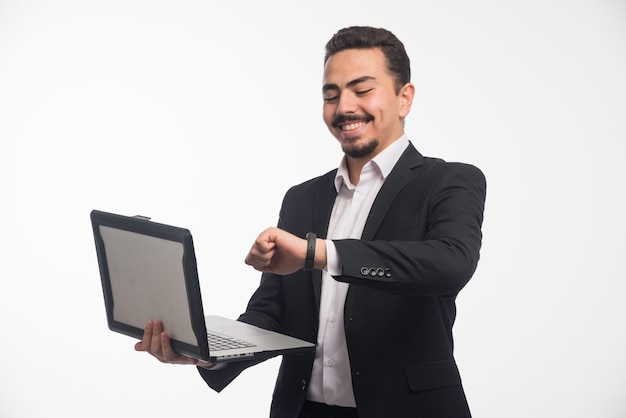 Un hombre de negocios en código de vestimenta sosteniendo una computadora portátil y señalando el tiempo.