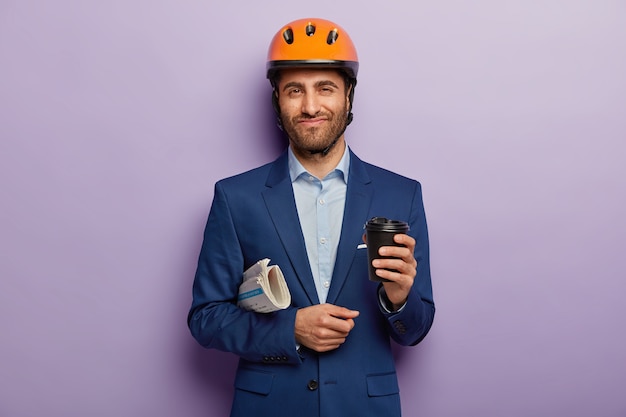 Hombre de negocios complacido posando en traje elegante y casco rojo en la oficina