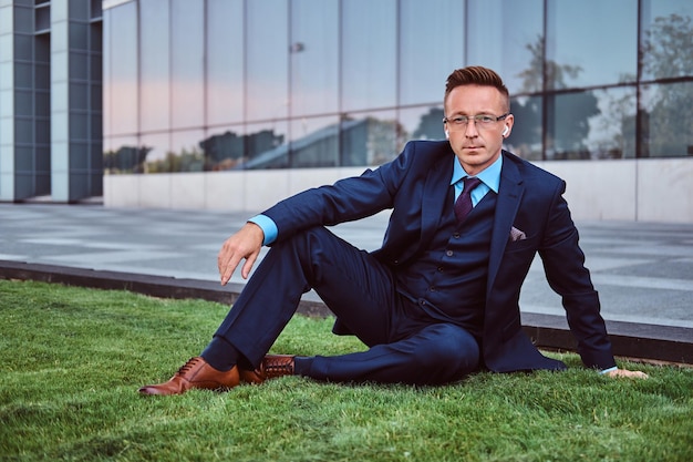 Hombre de negocios confiado vestido con un traje elegante sentado en un césped verde contra el fondo del paisaje urbano.