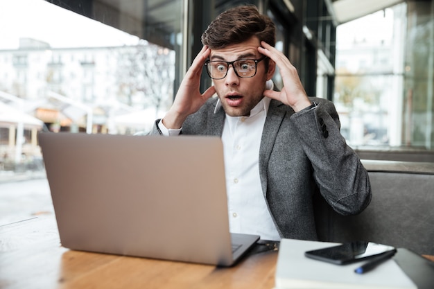 Hombre de negocios confundido preocupado en anteojos sentado junto a la mesa en el café mientras sostiene la cabeza y mira la computadora portátil