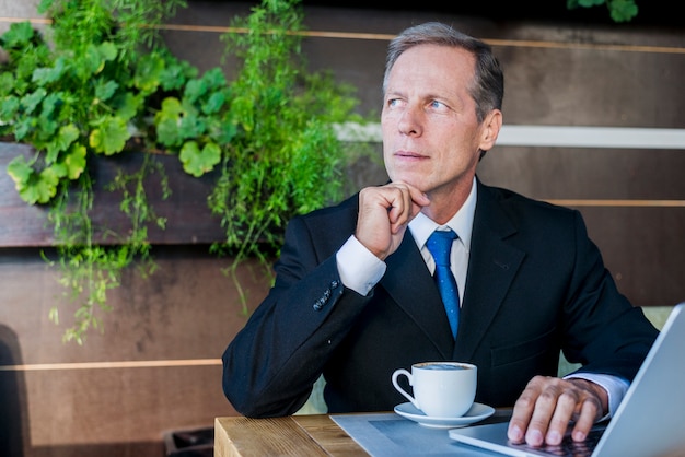 Foto gratuita hombre de negocios contemplado que se sienta en el café con la taza de café y el ordenador portátil en el escritorio