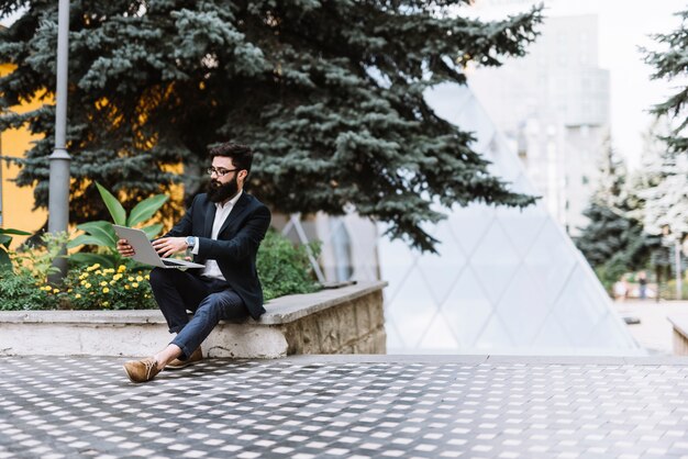 Hombre de negocios elegante joven que se sienta en campus usando el ordenador portátil