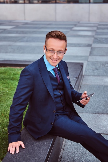 Un hombre de negocios elegante y sonriente vestido con un traje elegante sostiene un teléfono inteligente y mira hacia otro lado mientras se sienta al aire libre contra el fondo de un rascacielos.