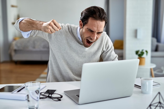 Hombre de negocios enojado gritando de frustración y a punto de golpear su computadora portátil después de leer malas noticias mientras trabajaba en casa