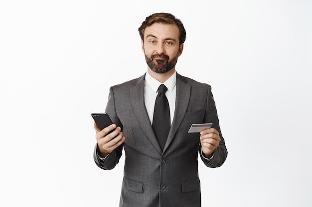 Hombre de negocios escéptico en traje haciendo muecas decepcionado sosteniendo la tarjeta de crédito y el teléfono móvil de pie sobre fondo blanco.