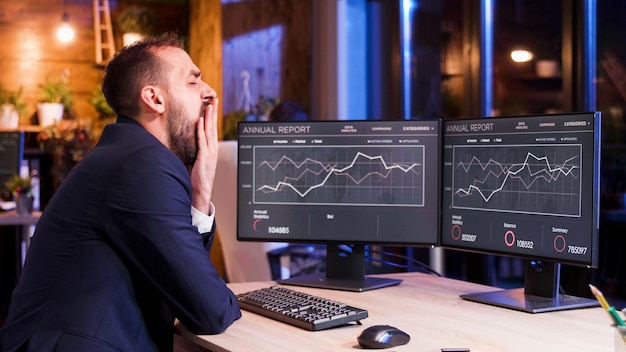 Hombre de negocios con exceso de trabajo bostezando en su escritorio de oficina a altas horas de la noche. La luz de la luna azul procedente de la ventana.