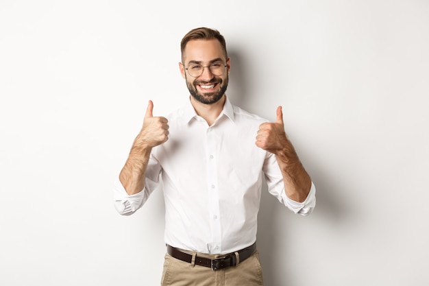 Hombre de negocios exitoso alabando el buen trabajo, mostrando los pulgares hacia arriba y sonriendo satisfecho, de pie