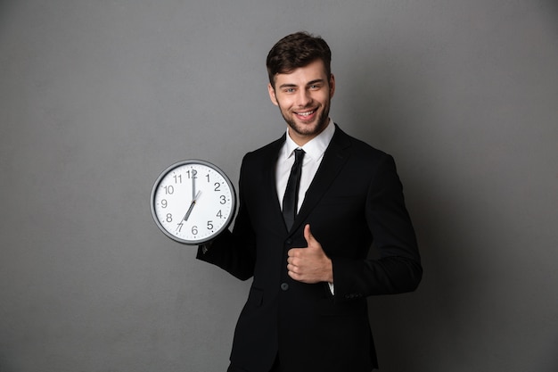 Hombre de negocios exitoso joven que sostiene el reloj mientras muestra el pulgar hacia arriba gesto y