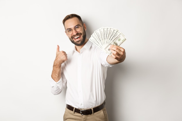 Hombre de negocios exitoso mostrando dinero dólares y pulgares arriba, sonriendo satisfecho, de pie