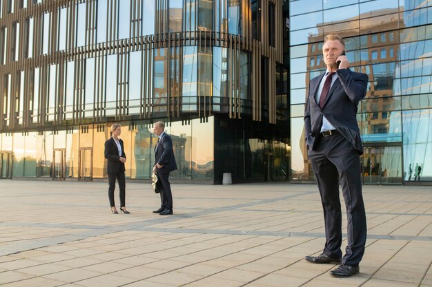 Hombre de negocios exitoso con traje de oficina, hablando por teléfono móvil al aire libre. Los empresarios y la fachada de cristal del edificio de la ciudad en segundo plano. Copie el espacio. Concepto de comunicación empresarial