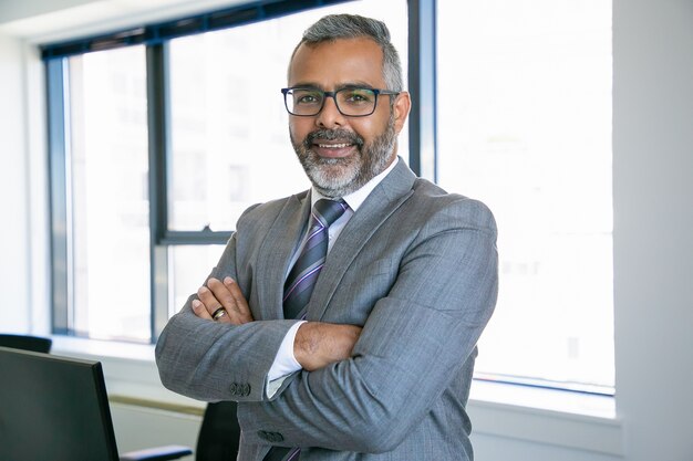 Hombre de negocios experimentado de pie en la sala de la oficina. Empleado de la oficina de contenido indio en anteojos sonriendo y posando con las manos juntas. Concepto de negocio, gestión y corporación