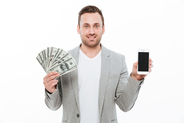 Hombre de negocios feliz alegre que sostiene el dinero y que muestra la exhibición del teléfono.