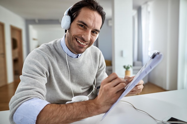 Hombre de negocios feliz con auriculares trabajando en papeleo en casa y mirando a la cámara