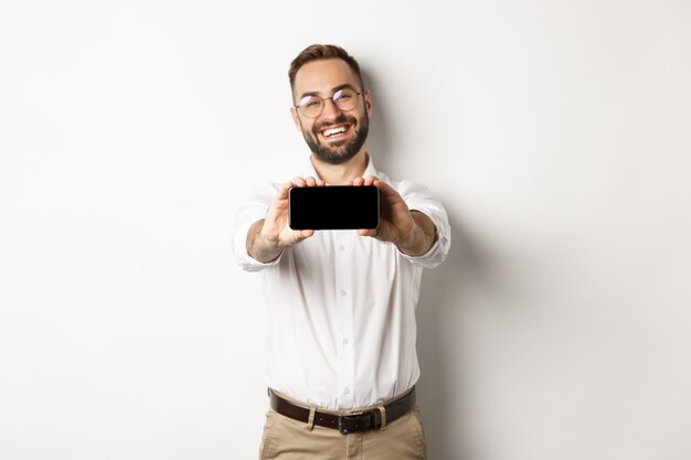 Hombre de negocios feliz mostrando la pantalla del móvil, sosteniendo el teléfono horizontalmente, de pie satisfecho