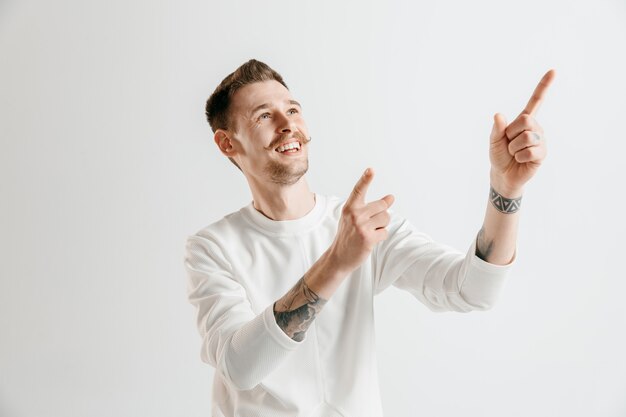 Hombre de negocios feliz de pie, sonriendo aislado sobre fondo gris de estudio. Hermoso retrato masculino de medio cuerpo. Los jóvenes satisfacen al hombre apuntando hacia arriba. Las emociones humanas, el concepto de expresión facial.