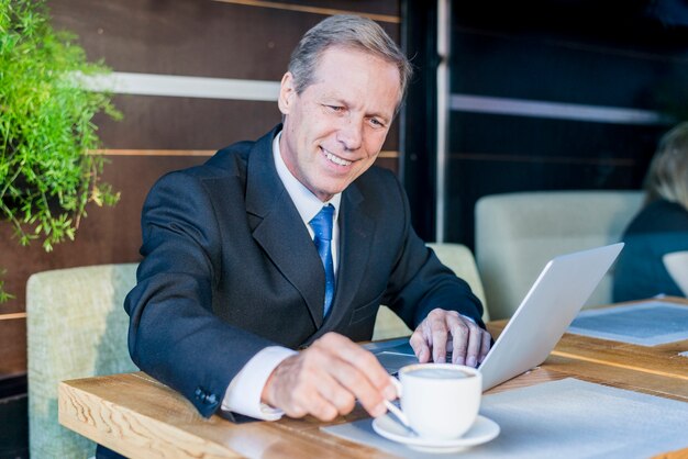 Hombre de negocios feliz que come la taza de café con el ordenador portátil en el escritorio en caf�