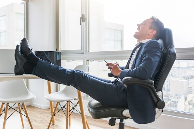 Foto gratuita hombre de negocios feliz que se sienta en silla en el lugar de trabajo usando móvil