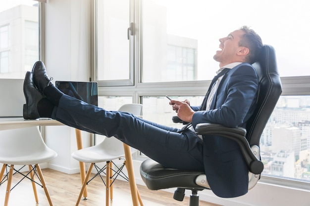 Hombre de negocios feliz que se sienta en silla en el lugar de trabajo usando móvil