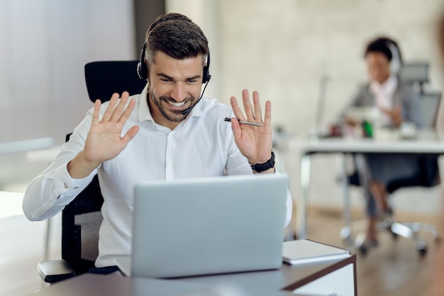 Un hombre de negocios feliz saludando a alguien mientras hace una videollamada a través de una laptop en la oficina