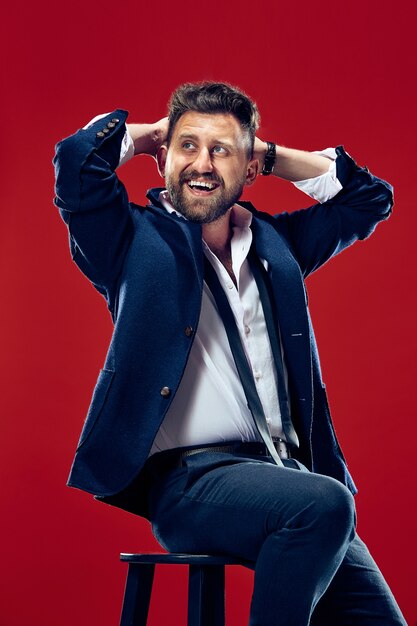 Hombre de negocios feliz sentado y sonriendo aislado sobre fondo rojo de estudio.