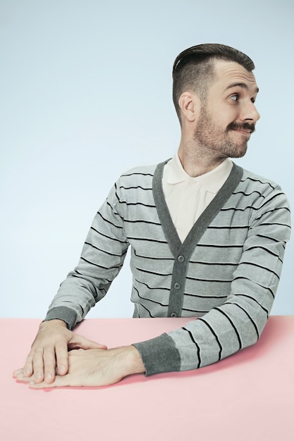 El hombre de negocios feliz y sonriente sentado a la mesa sobre fondo azul de estudio. El retrato en estilo minimalista